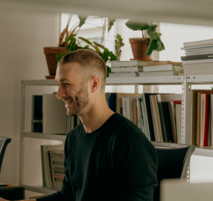 A smiling man with short hair sitting at a desk in an office with shelves and plants in the background